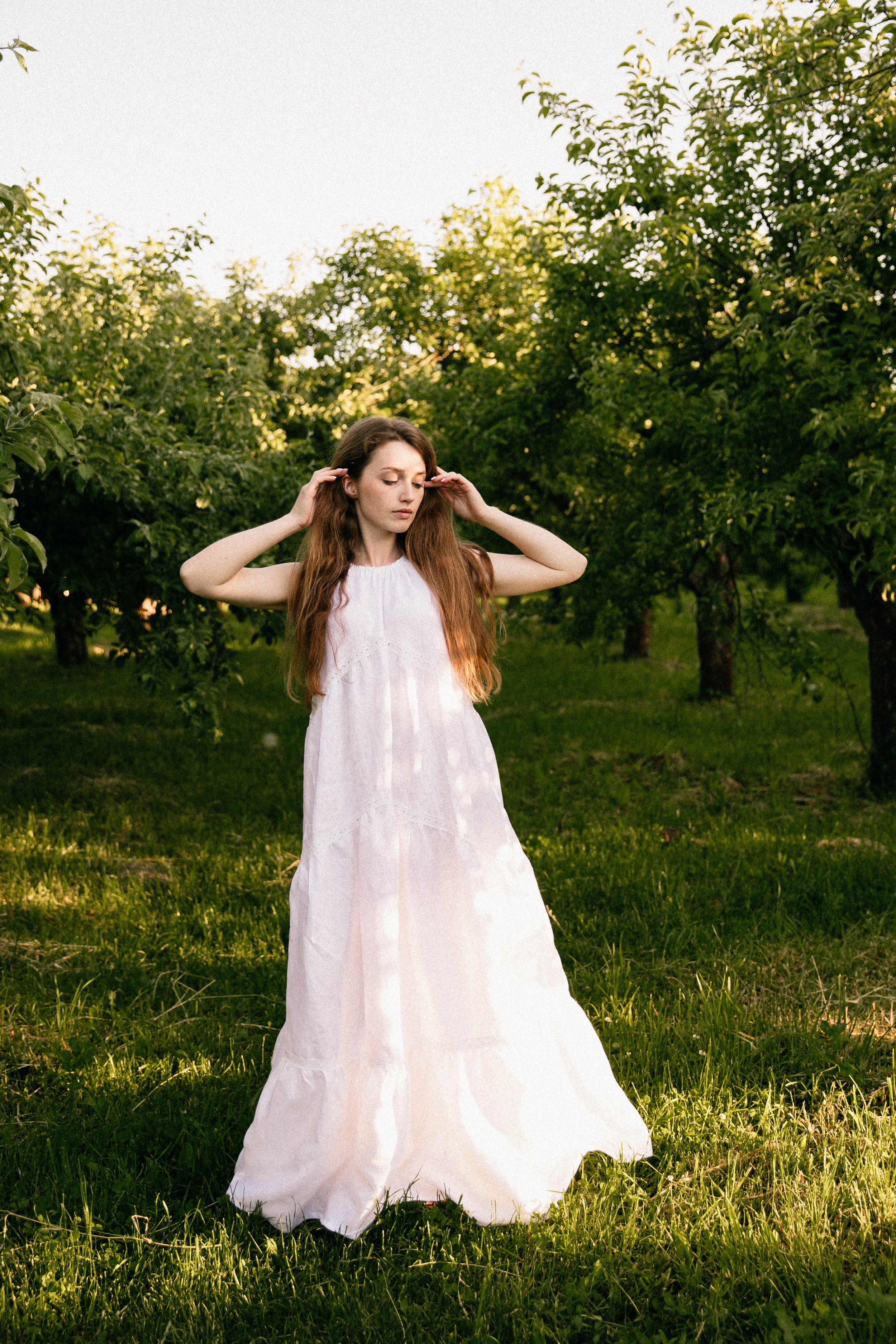 Woman in a white linen wedding dress standing in an orchard with trees and grass around her.