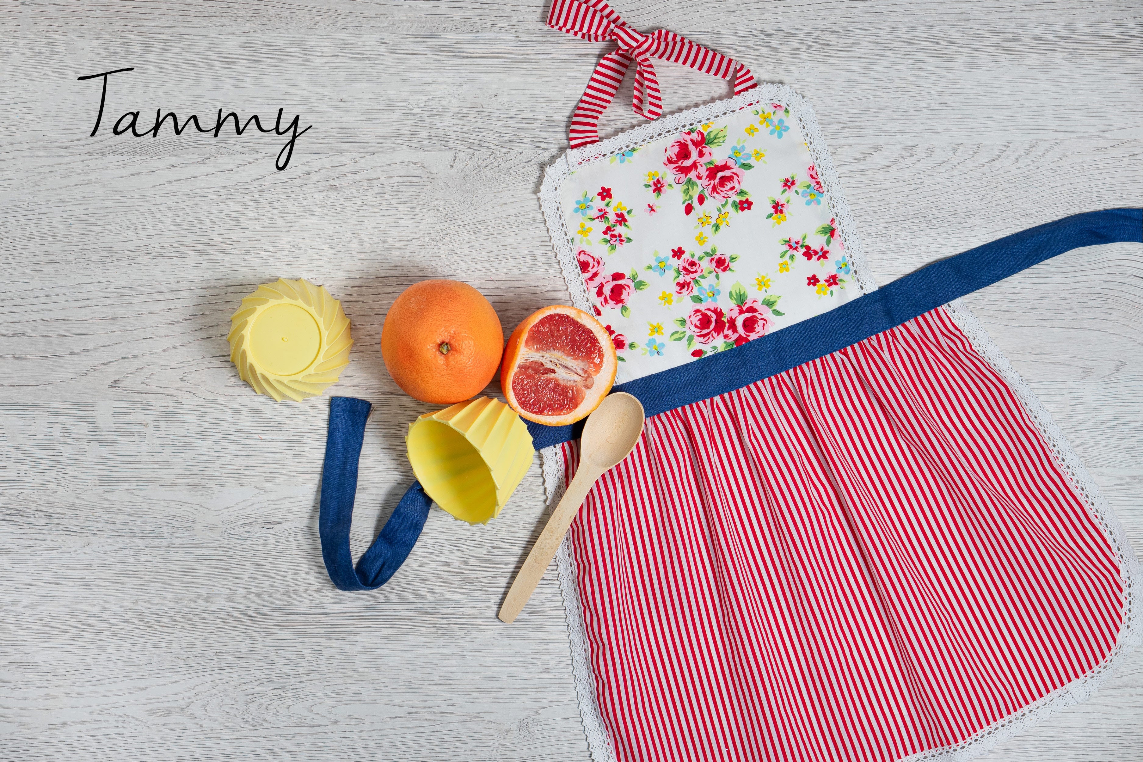 Floral cotton apron for girls with red and white pattern on a light wooden surface with colorful items.