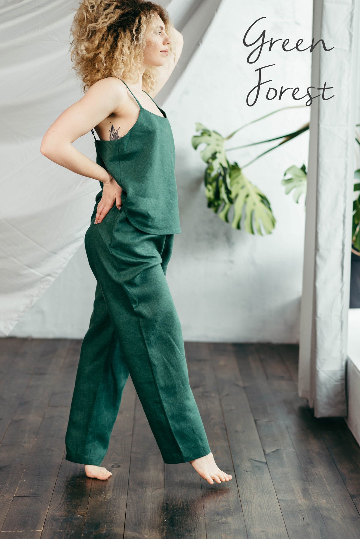 Woman wearing a green linen pyjama top and bottoms standing in a room with white curtains and a plant.