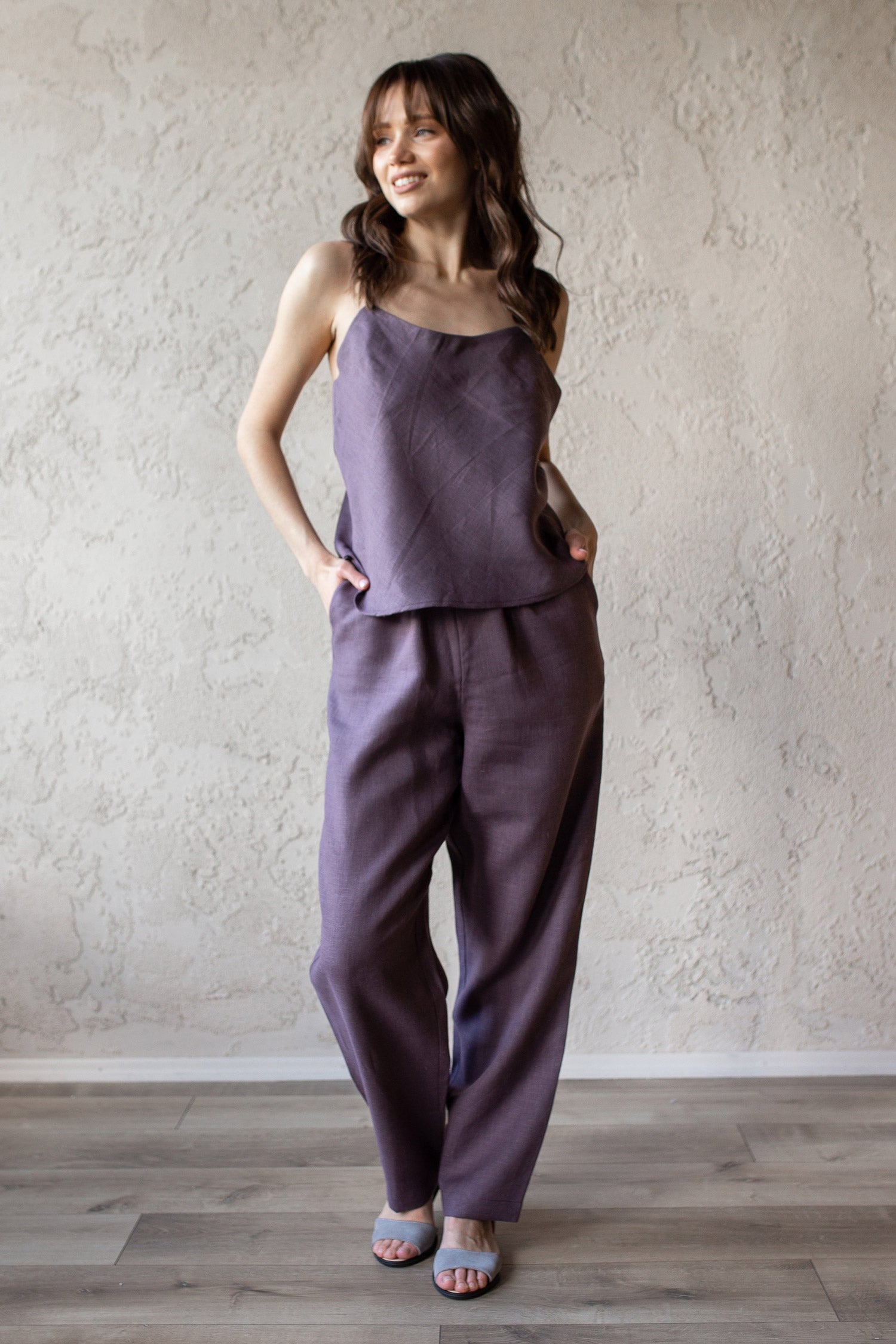 Woman wearing a purple pajama top and pants standing against a plain wall.
