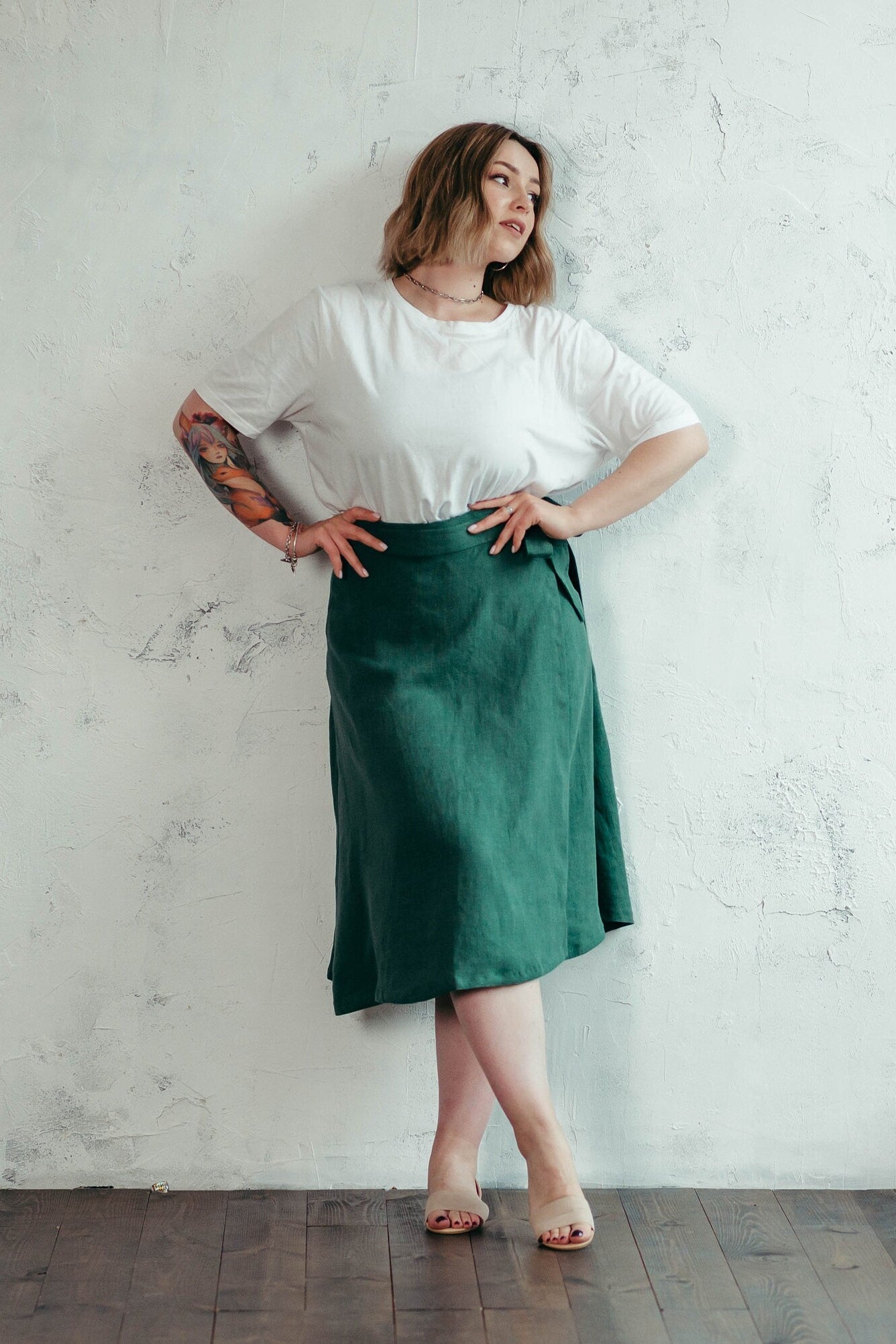 Woman wearing a white top and green linen midi wrap skirt standing against a white wall.