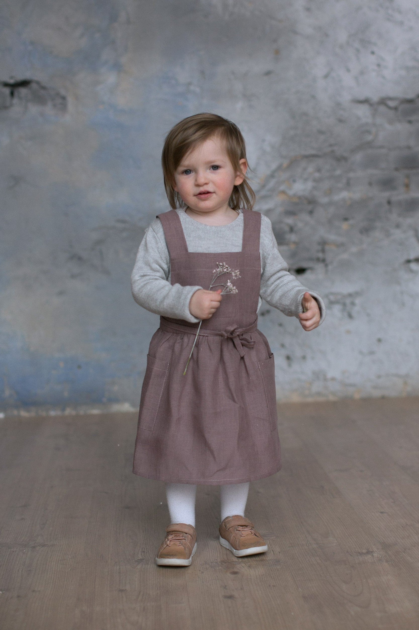 Toddler girl is wearing a brown dress apron while holding a flower