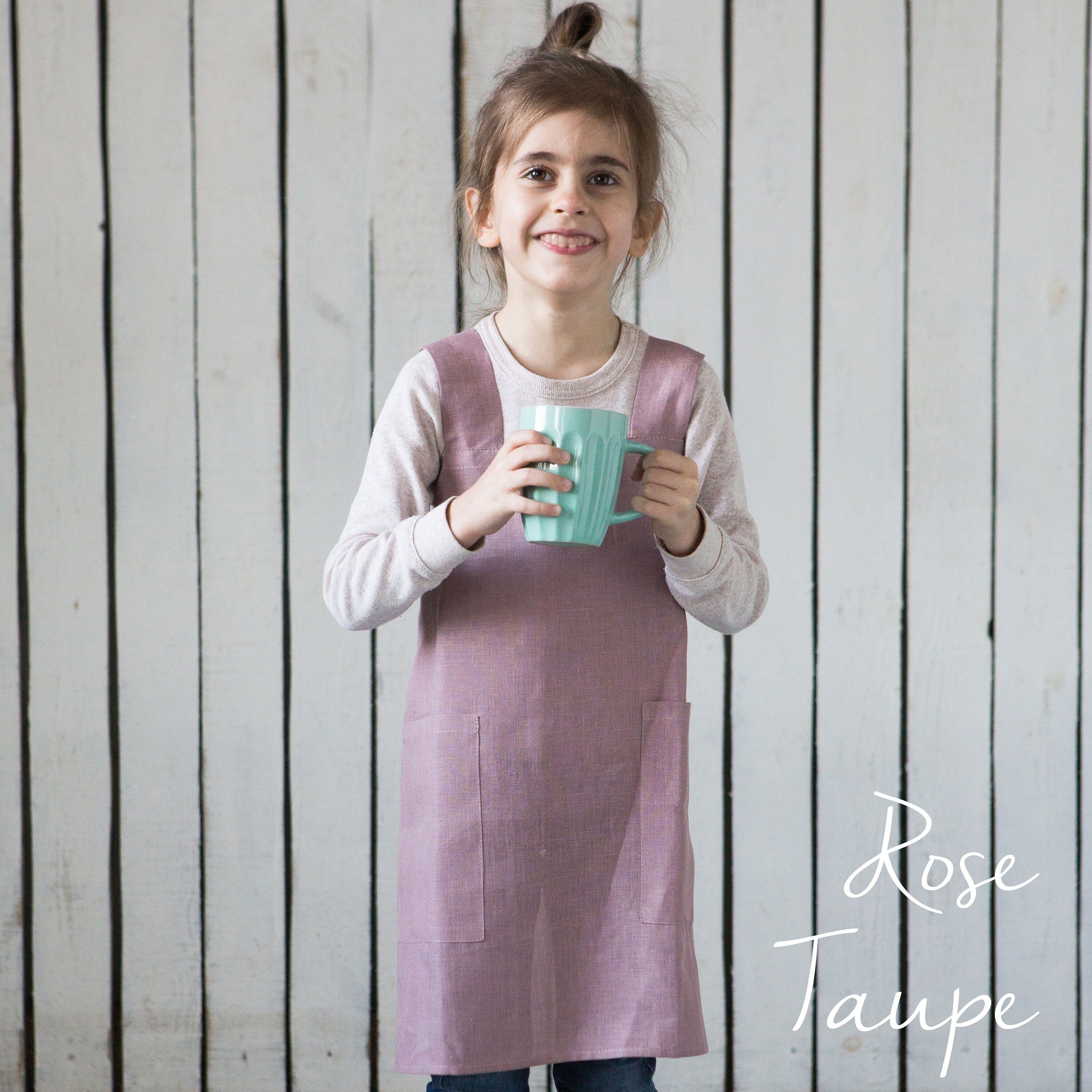 Child wearing a rose taupe linen smock apron holding a mug against a wooden background