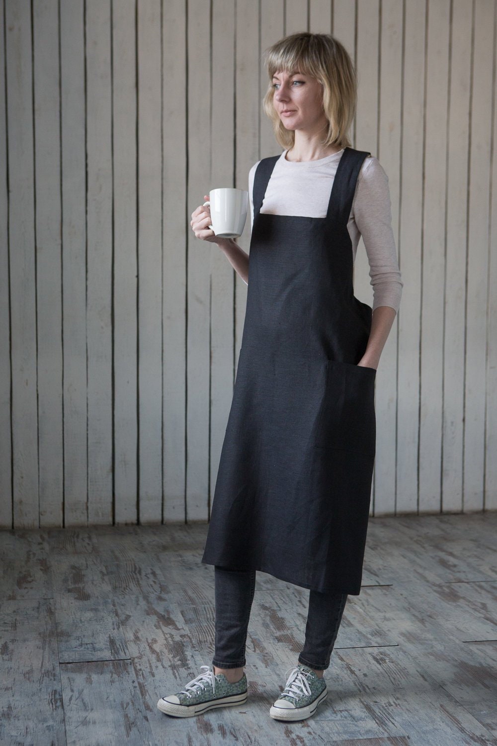 Woman wearing a black inen apron holding a white mug against a wooden wall.