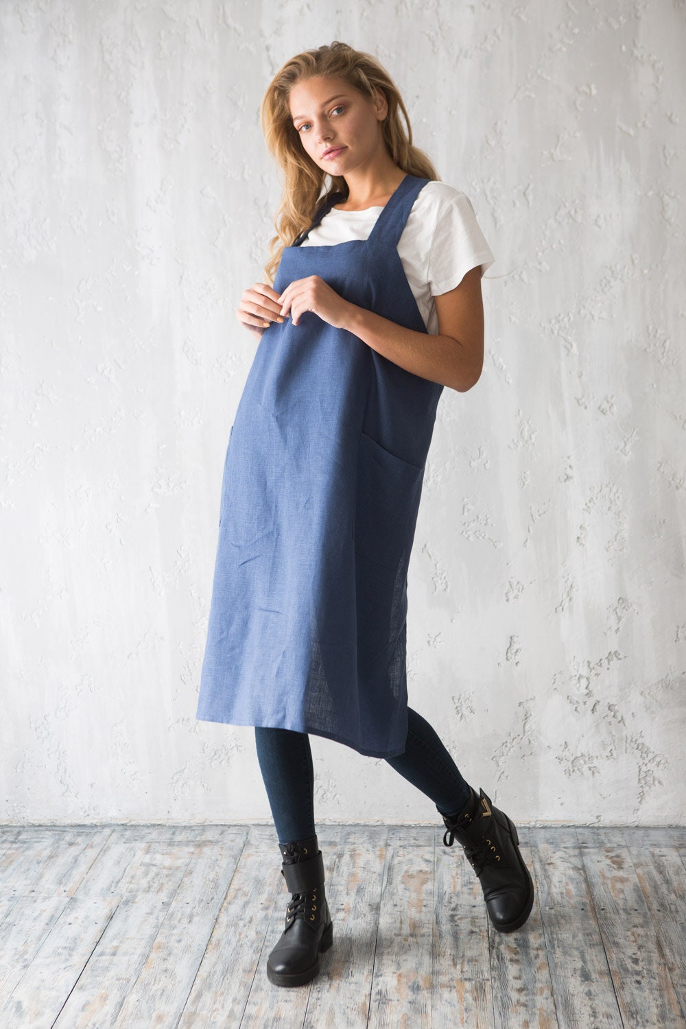 Woman wearing a blue linen smock apron over a white shirt against a light gray wall.