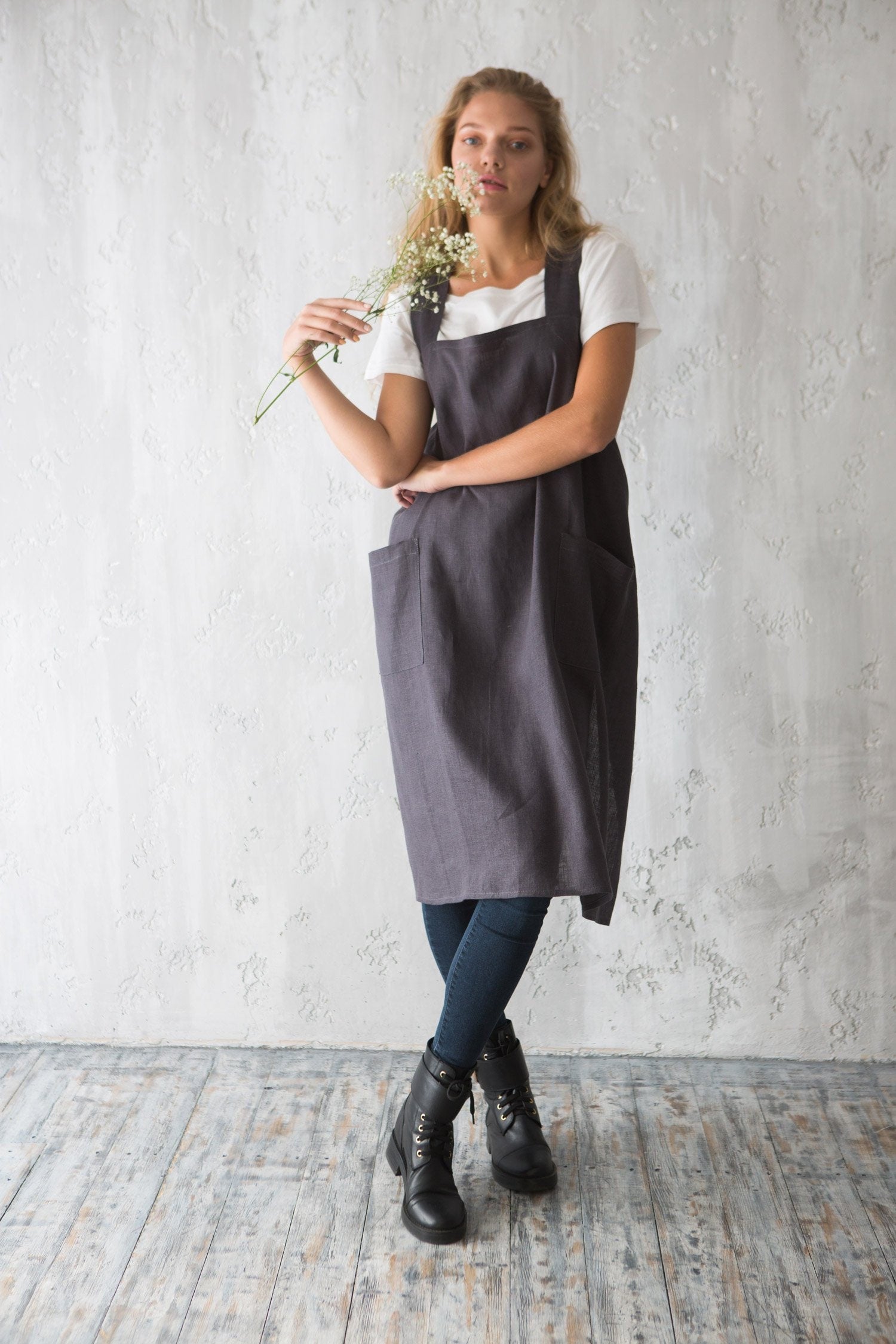 Woman wearing a dark grey linen apron over a white shirt, holding flowers against a light gray wall.