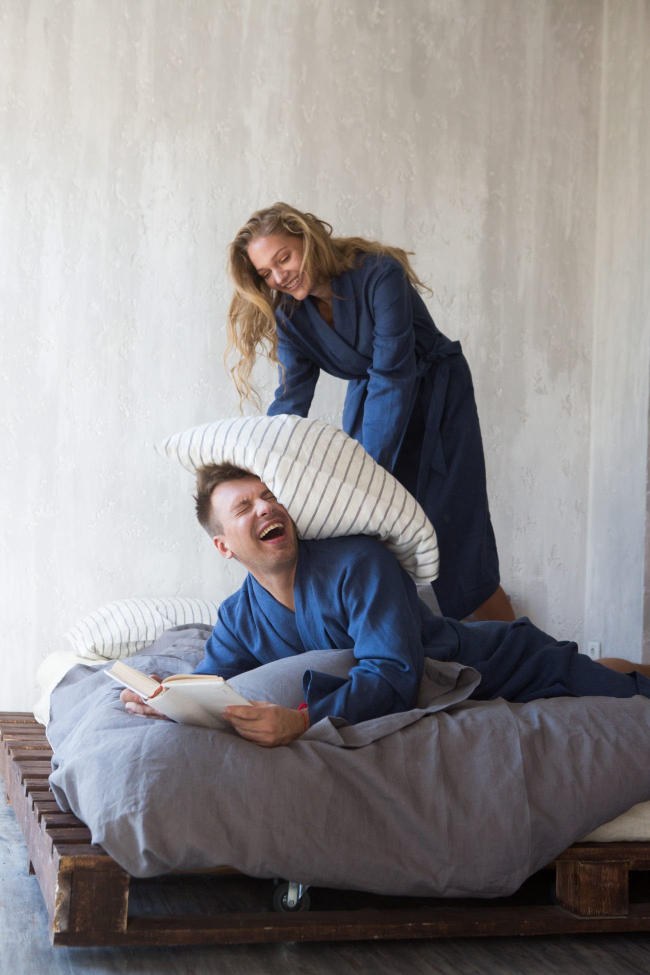 Man lying on a bed reading a book with a woman standing behind him, both wearing matching linen robes.