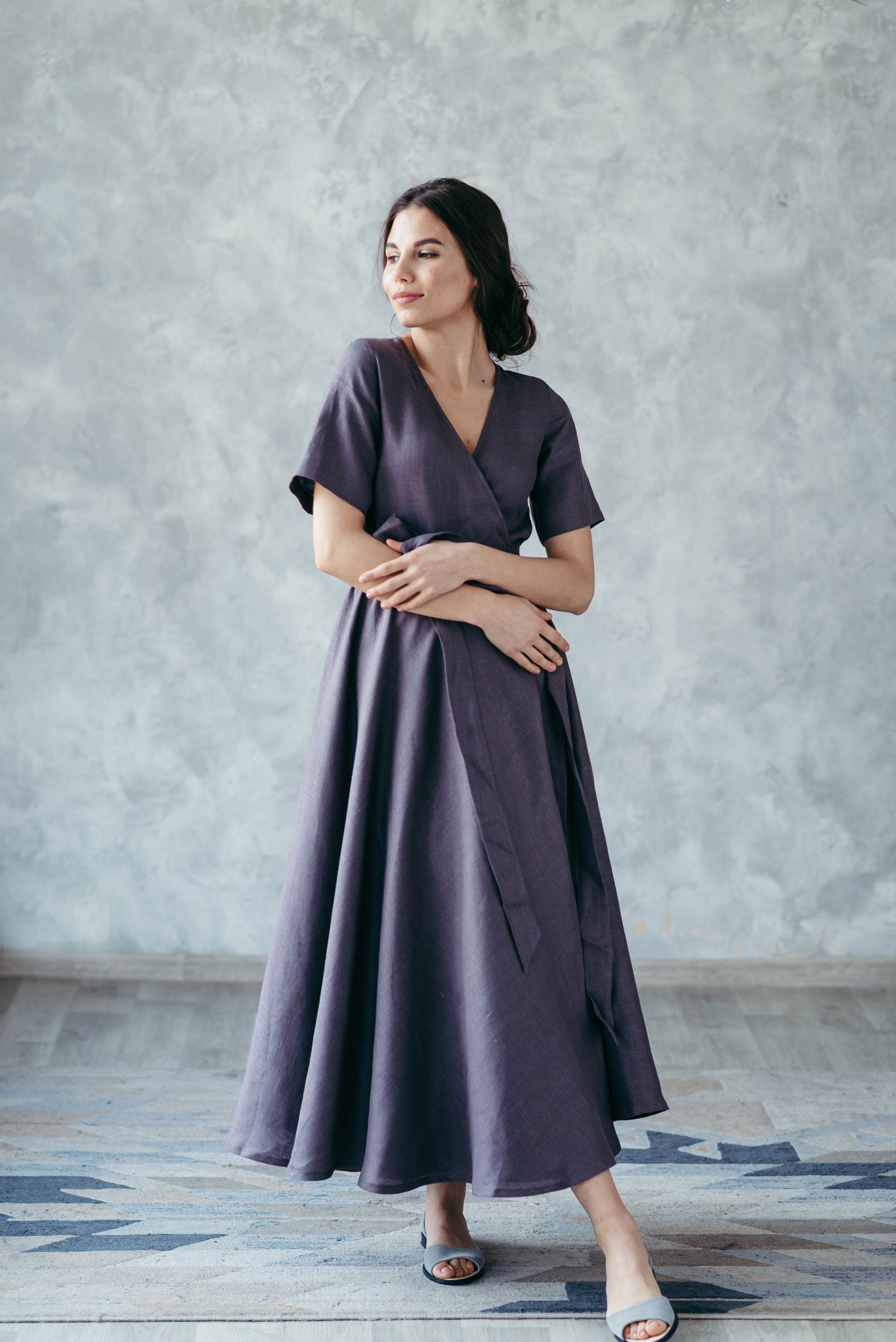 Woman wearing a long, dark purple linen wrap dress standing against a plain wall.