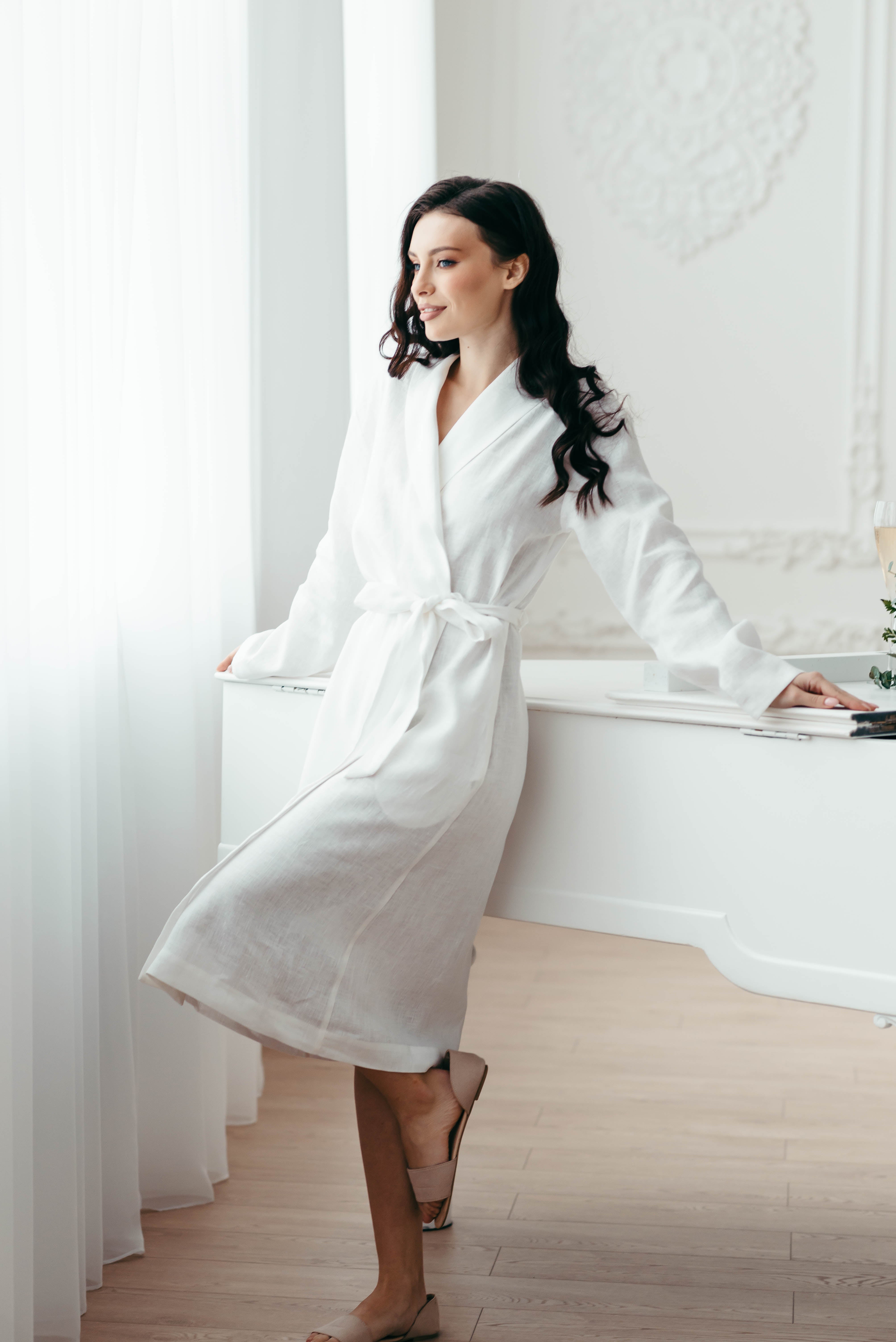 Woman in a white robe standing in a bright room with large windows.