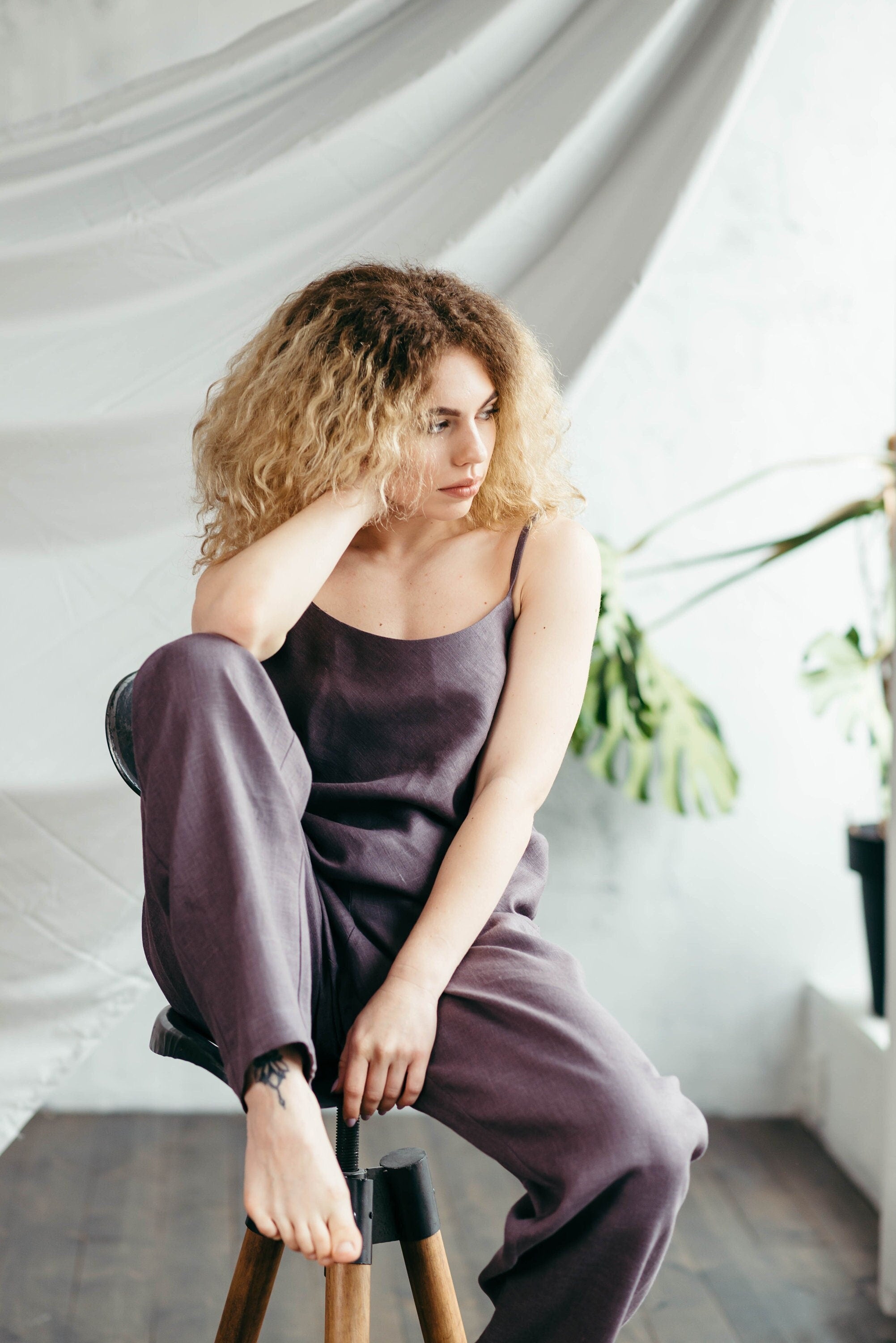 Woman in a purple pyjamas set sitting on a stool with a white curtain and plant in the background