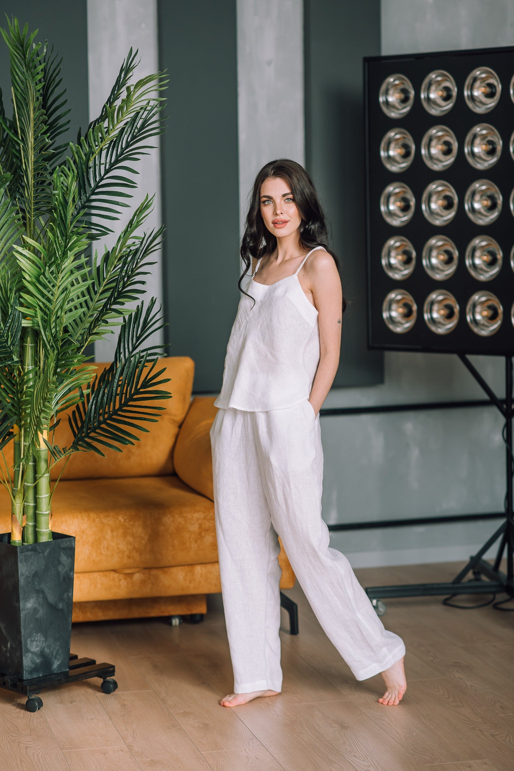 Woman in a washed white pyjamas standing in a room with a plant and a couch.