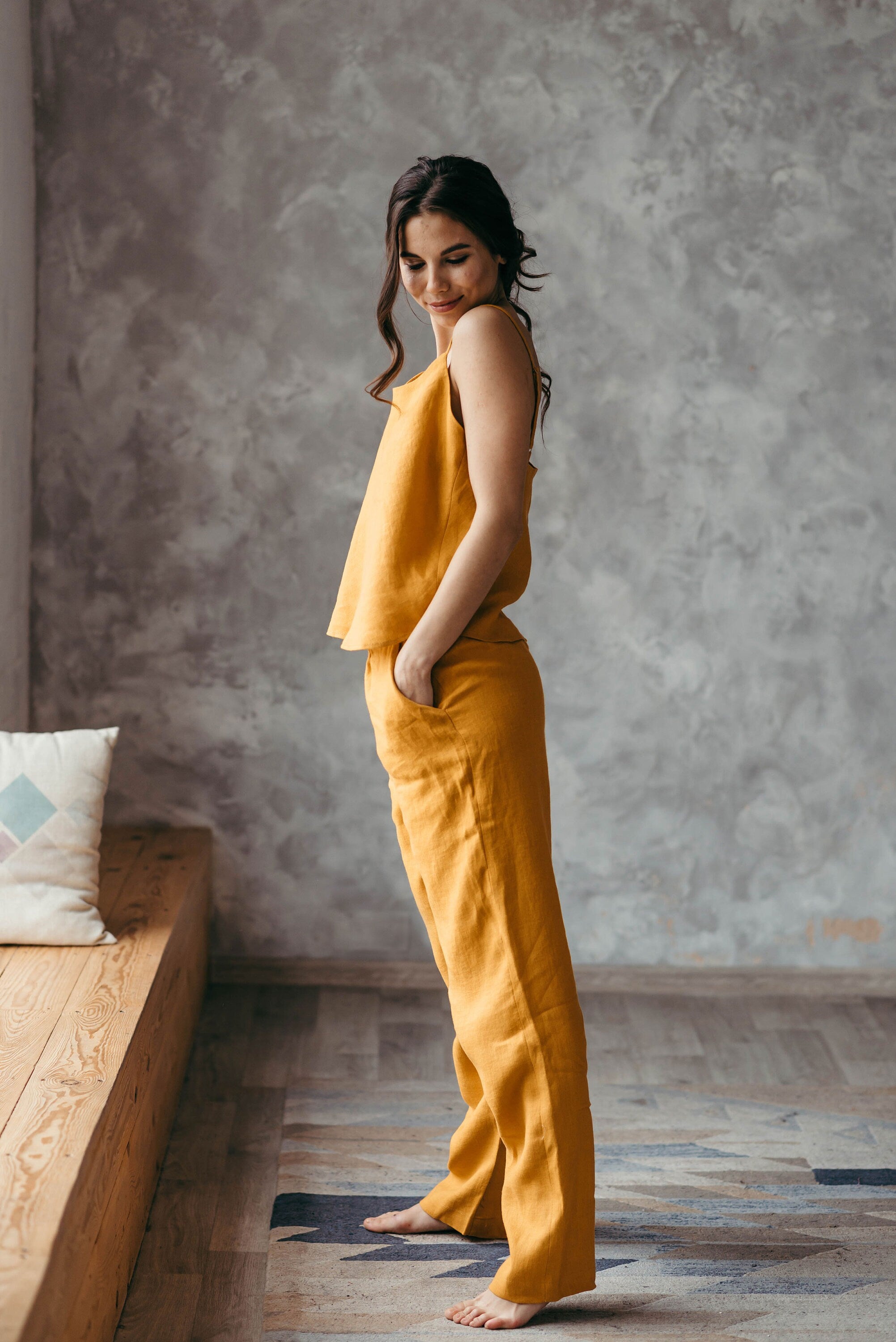 Woman in a yellow ochre washed linen pyjamas set standing in a room with a textured wall and wooden bench.