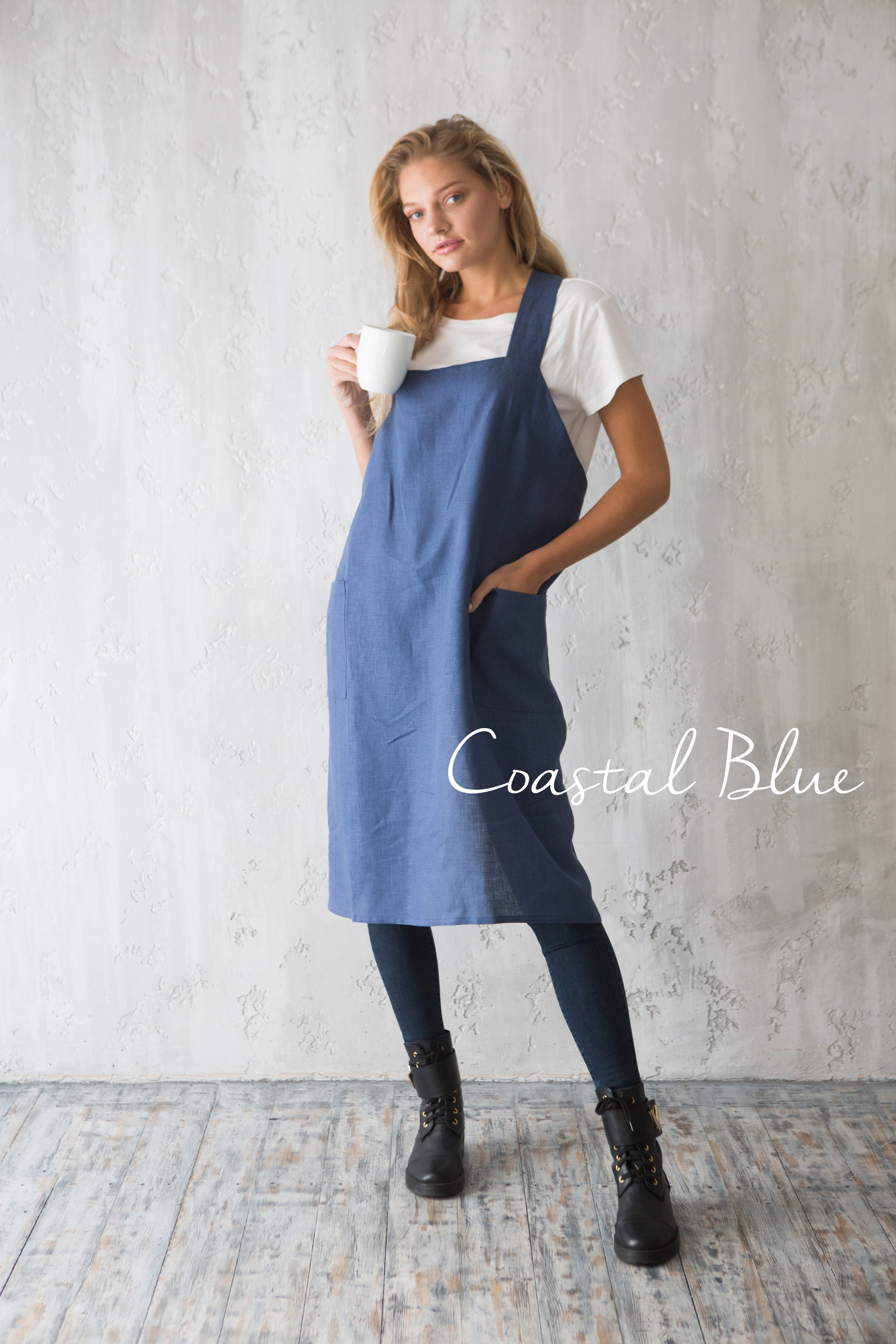 Woman wearing mid blue linen pinafore apron and drinking coffee.