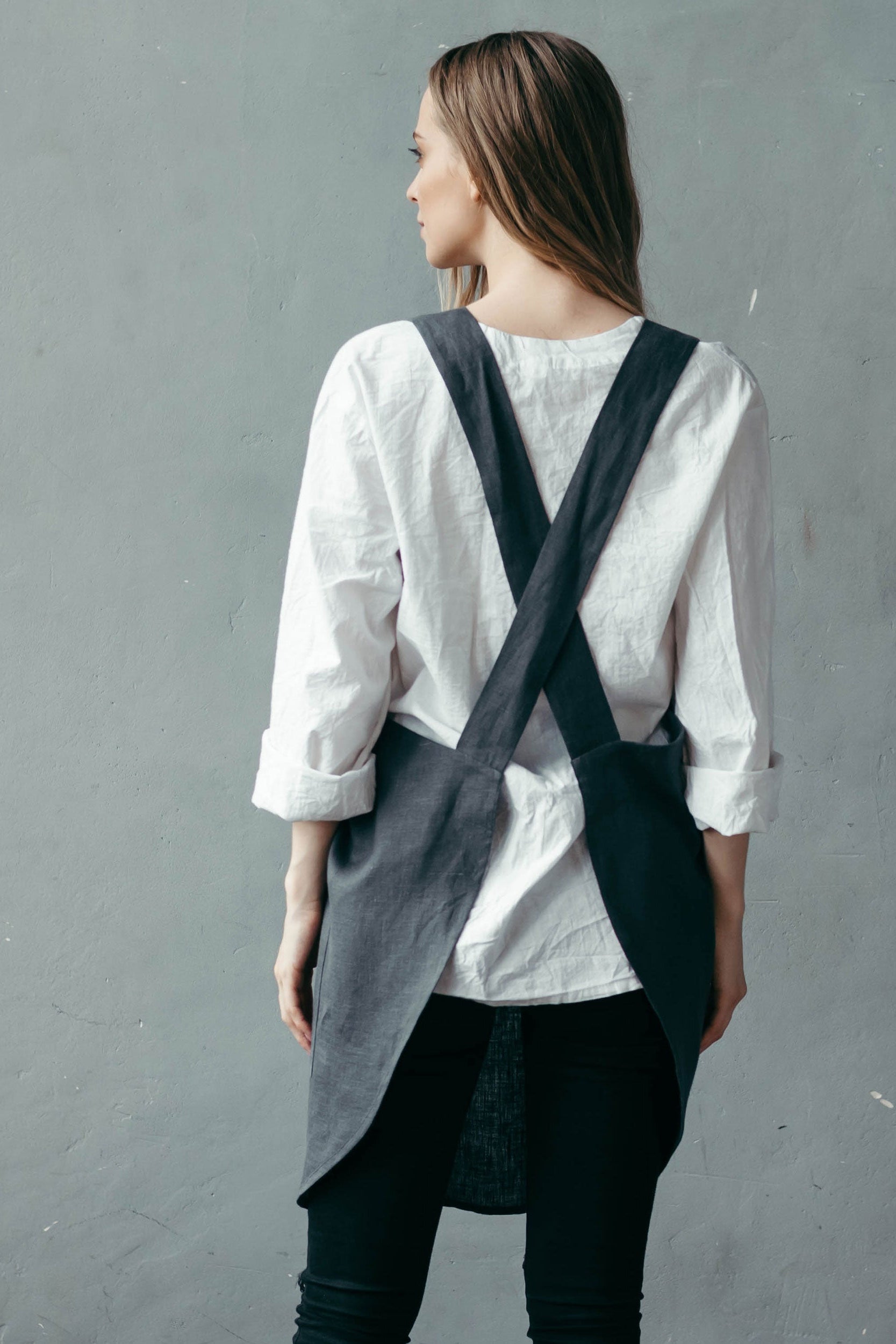 Person wearing a white shirt and dark linen apron overalls against a gray background