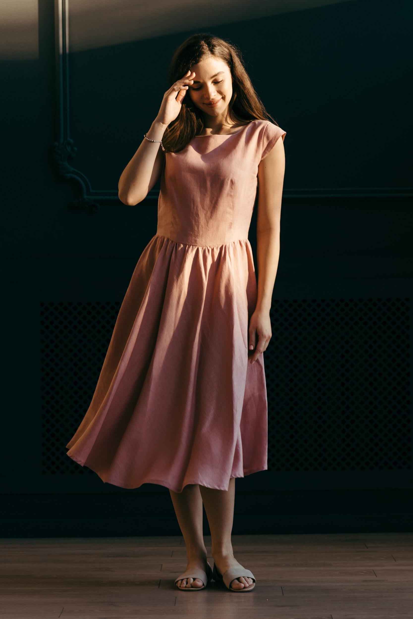 Woman in a pink midi linen dress standing against a dark wall.