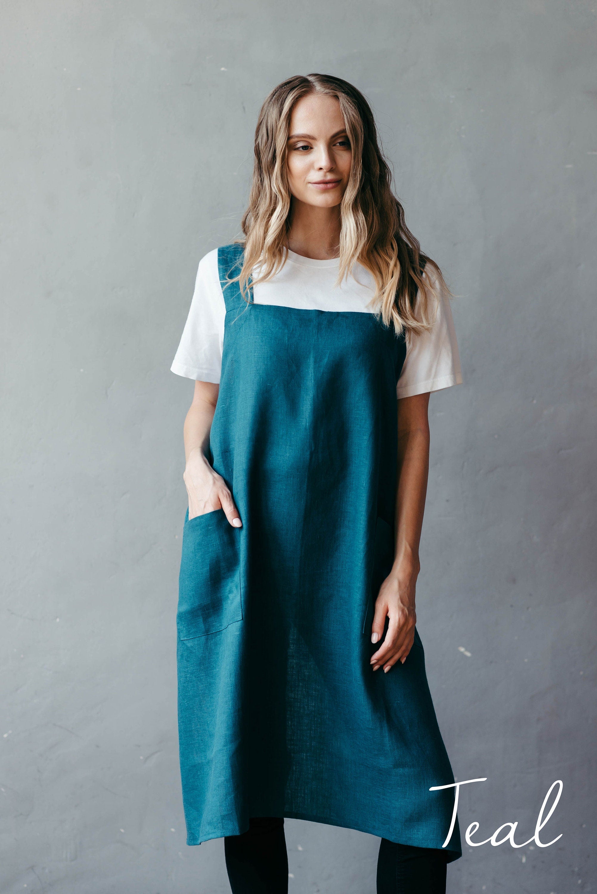 Woman wearing a teal linen smock apron over a white shirt against a gray background