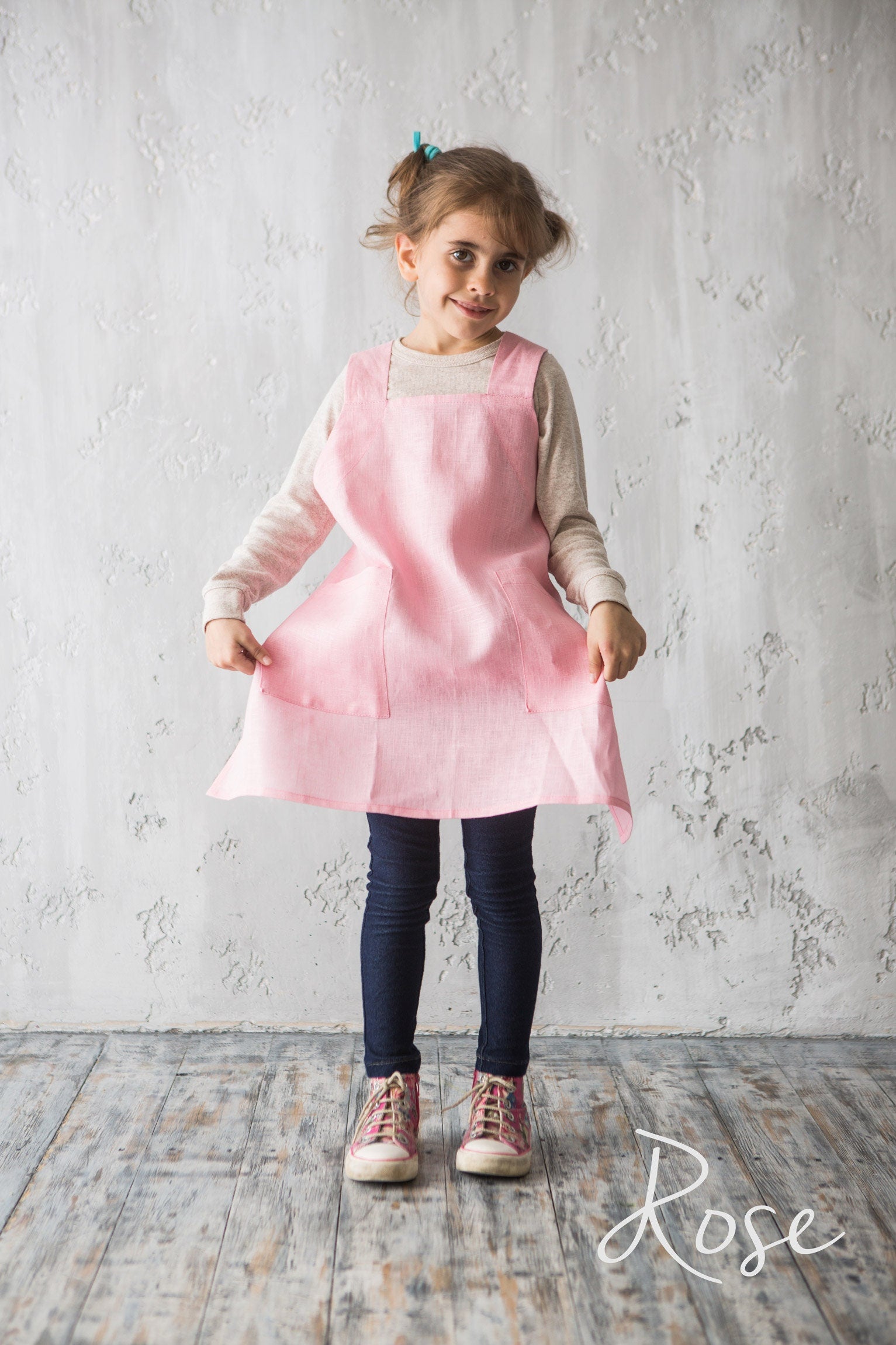 Young girl wearing a pink crossover linen apron standing against a textured white wall.