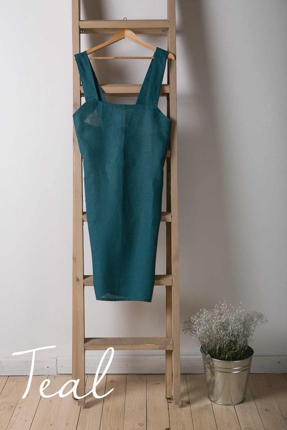 Teal linen apron hanging on a wooden ladder against a white wall with a plant in the corner.