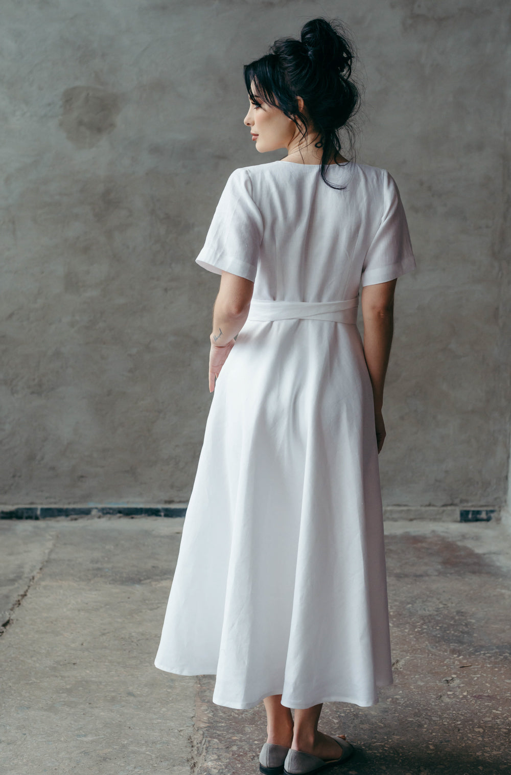 Woman wearing a white linen dress standing against a plain wall.