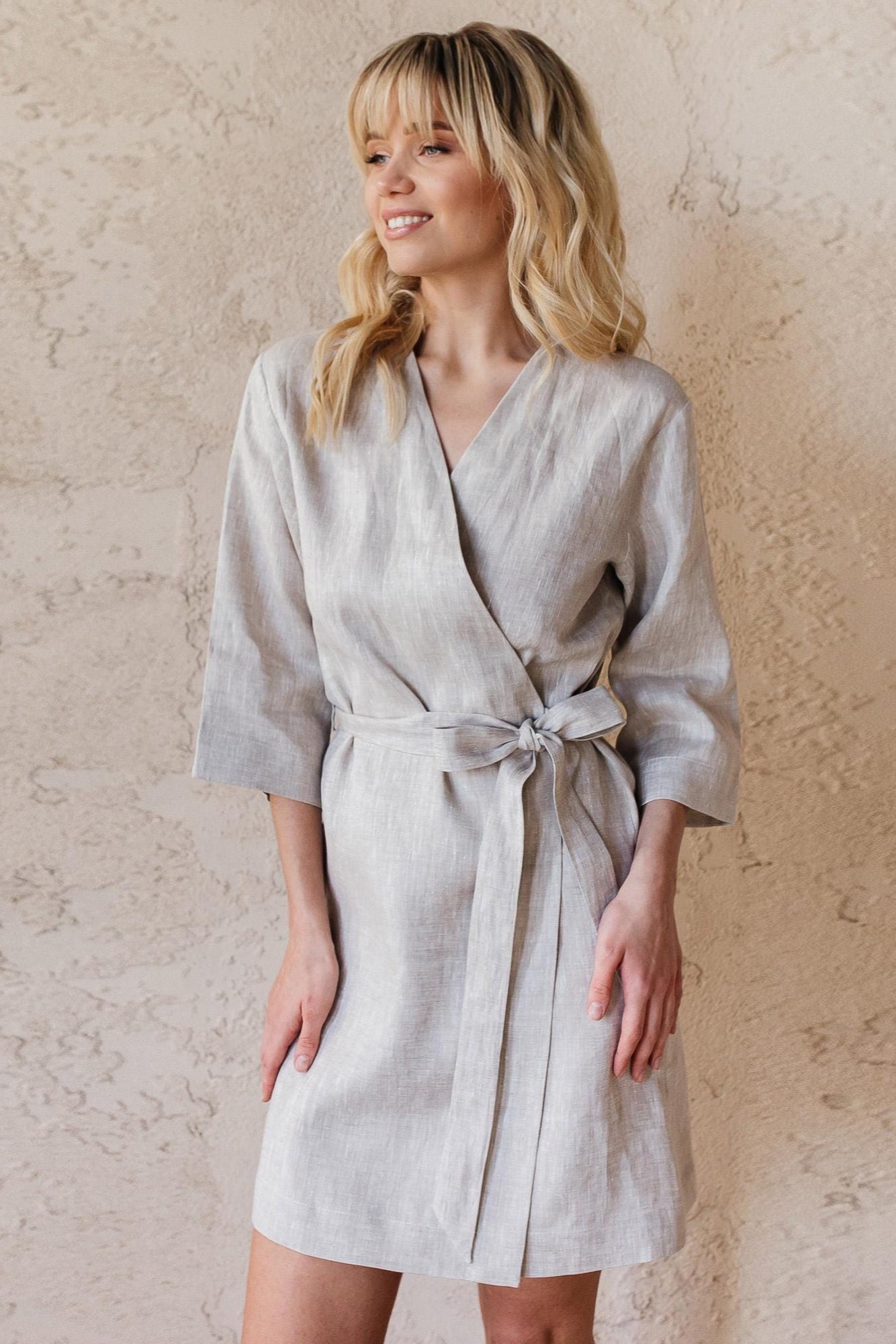A woman wearing a natural gray short linen robe against a beige wall