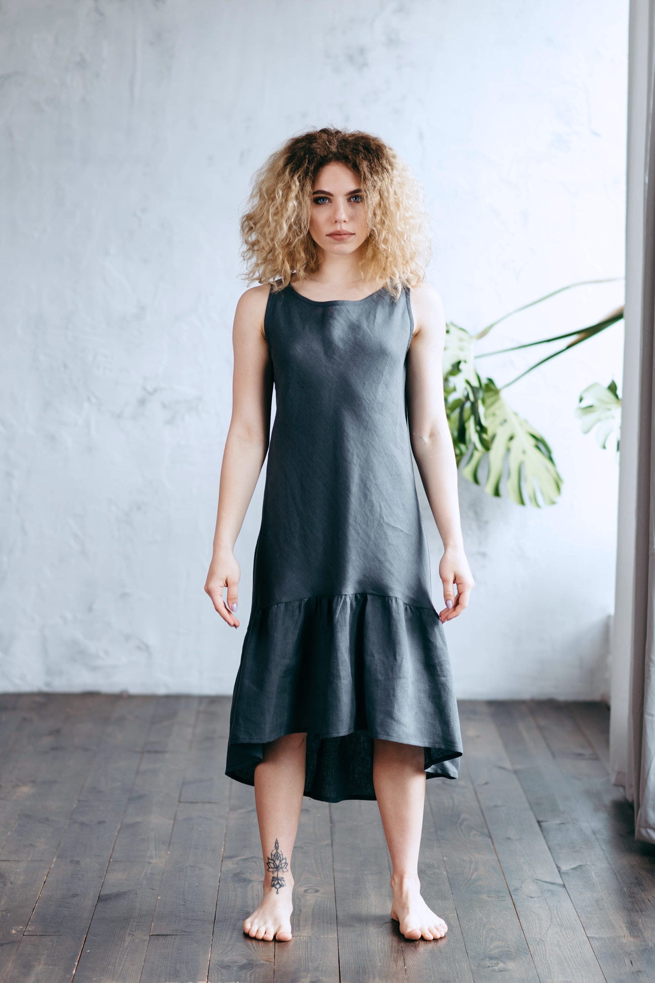 Woman wearing a dark gray linen dress standing in a room with a white wall and plant.