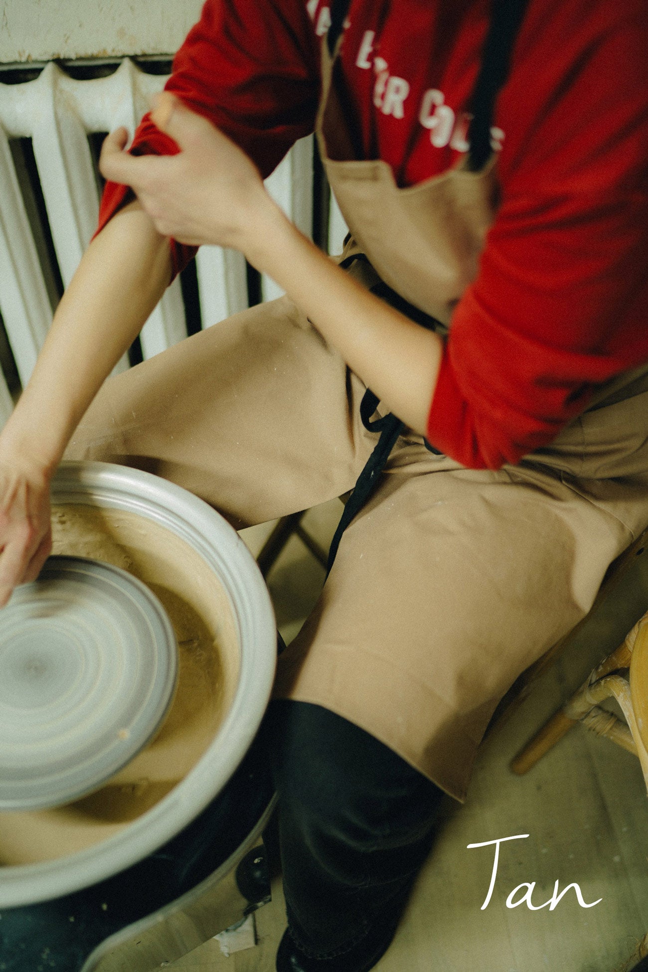 Potter working with clay on a potter's wheel, wearing a red shirt and tan pottery apron.