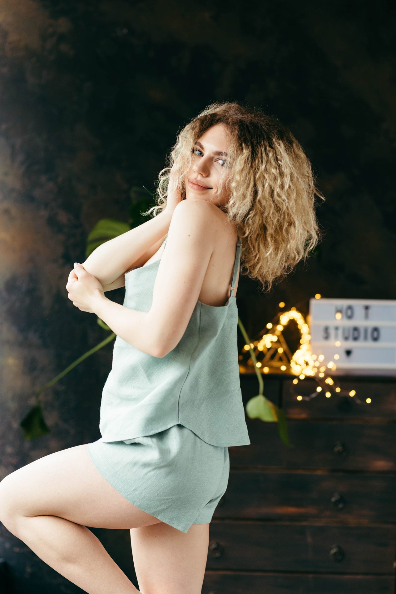 Woman in a light green natural linen pyjamas posing in front of a dark background.
