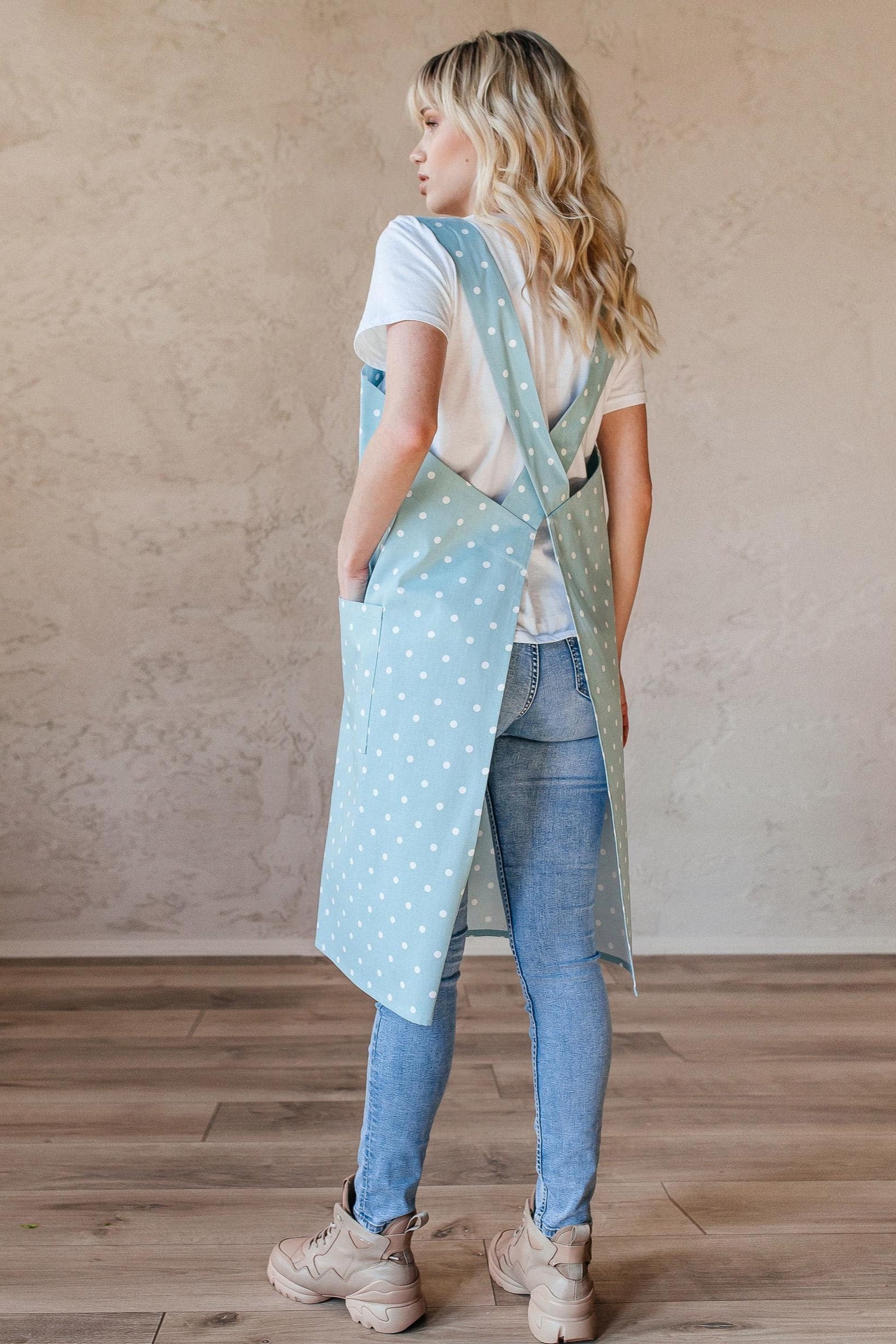 Woman wearing a light blue polka dot pinafore apron over a white shirt and jeans in a room with wooden flooring and a beige wall.