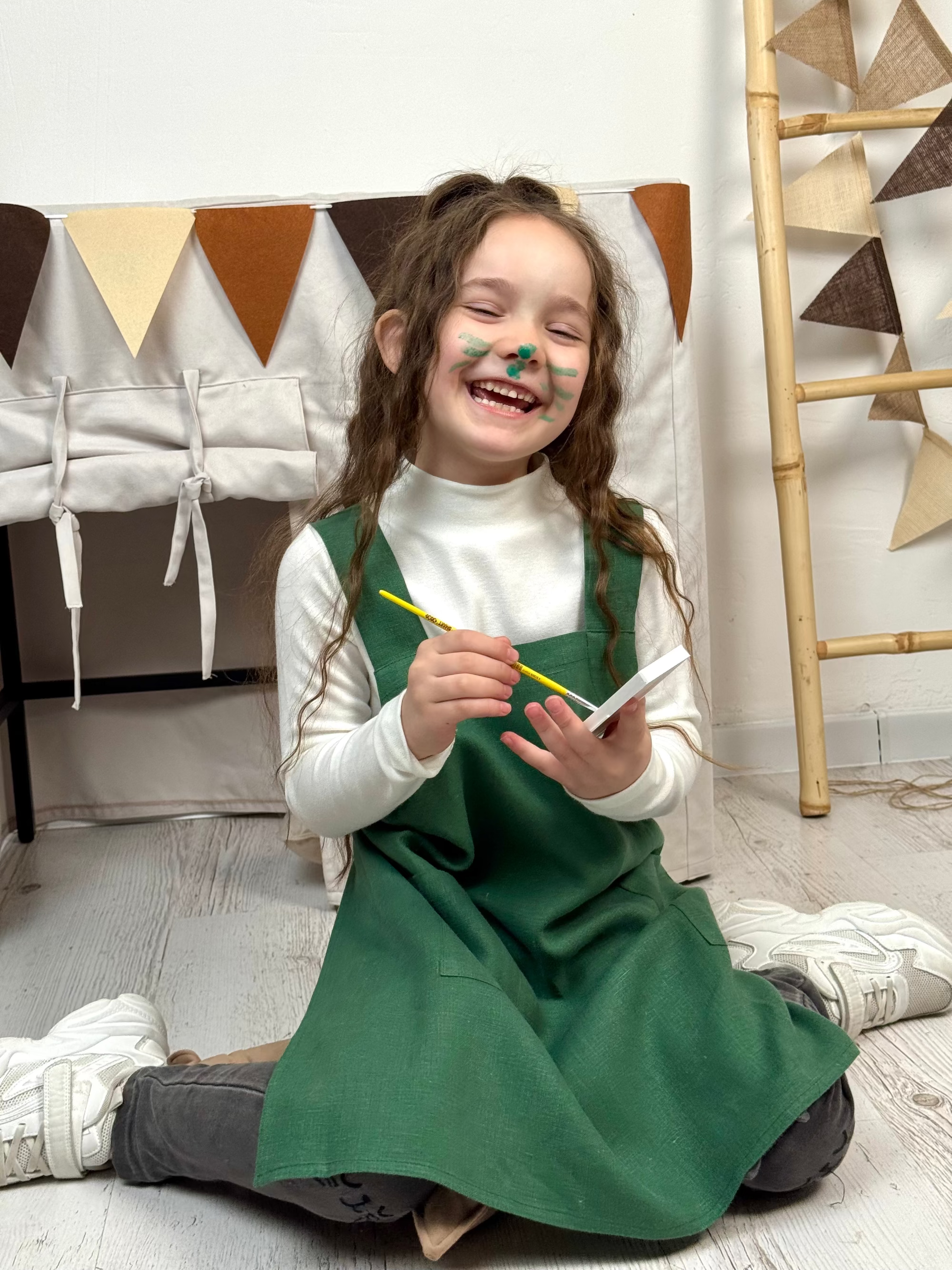 Child in a green linen pinafore apron a yellow paintbrush while painting a picture in her playroom.