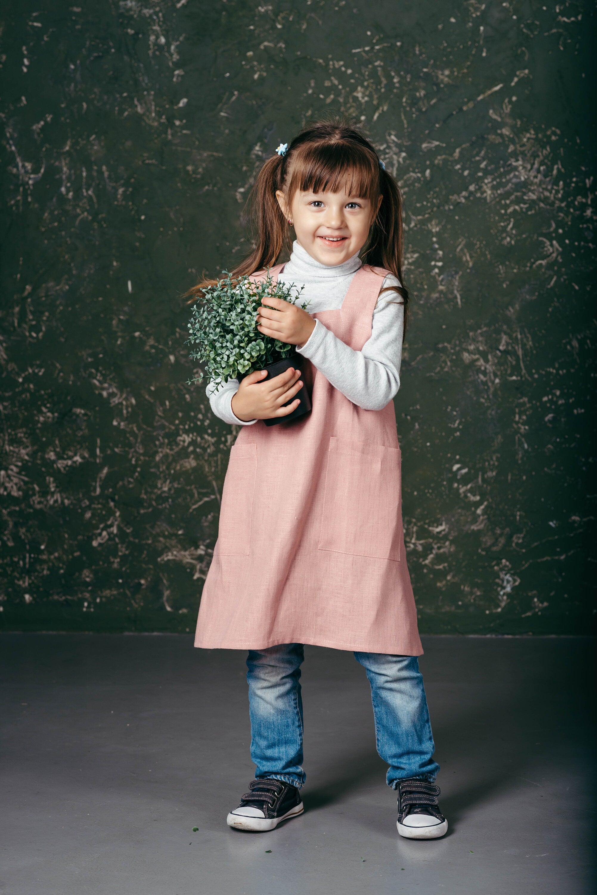 Tpddler girl in a pink linen pinafore linen apron for kids is holding a plant agains