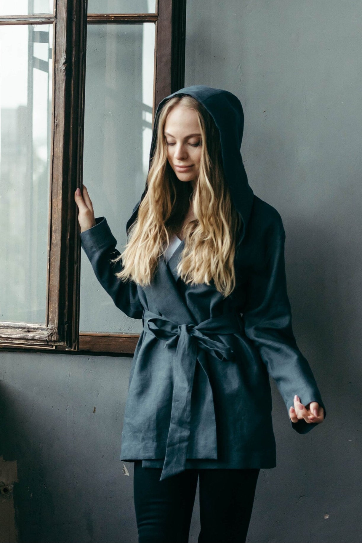 Woman in a dark grey linen jacket standing by a window in a room with gray walls.