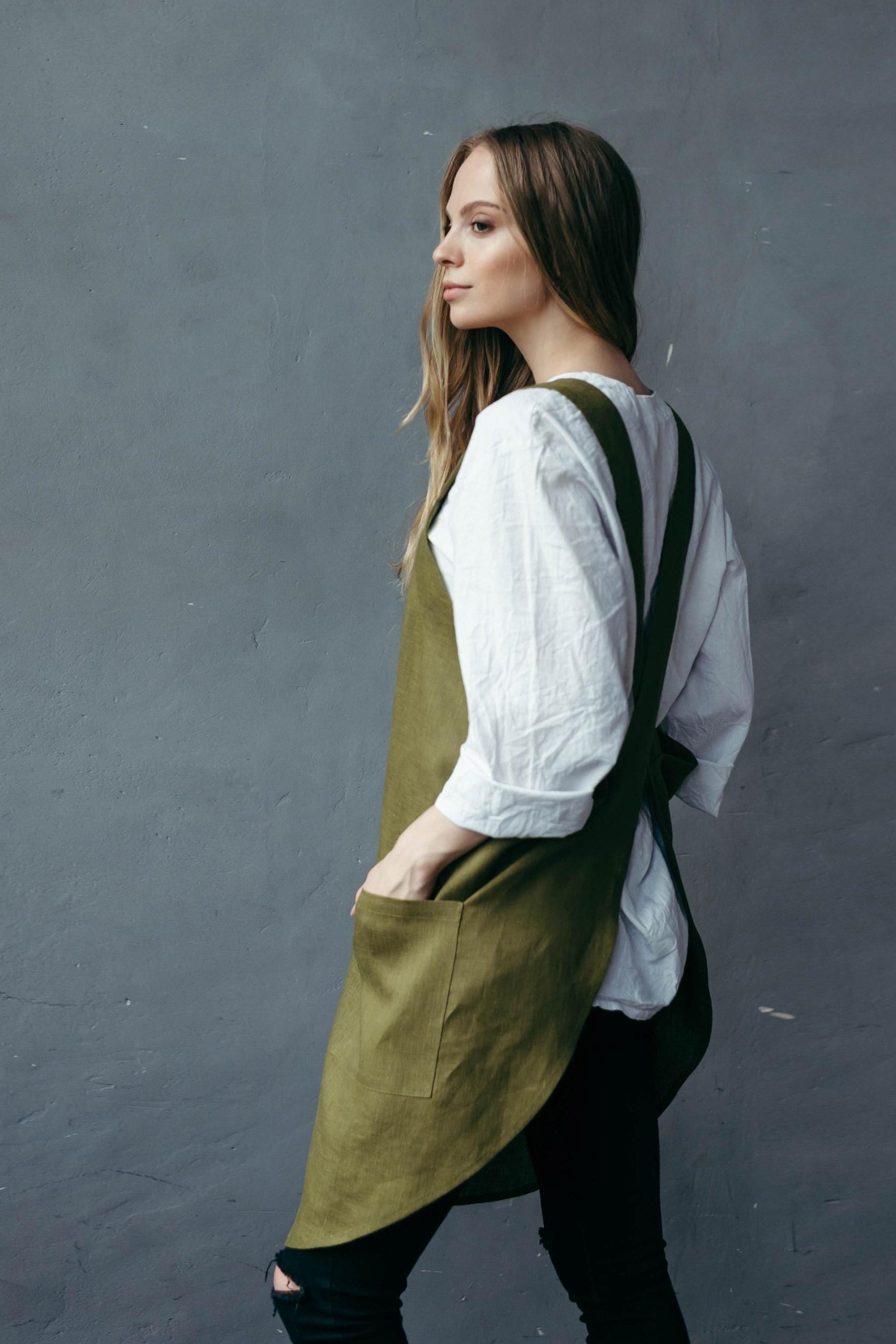 Woman wearing a khaki green linen smock apron over a white shirt against a dark background