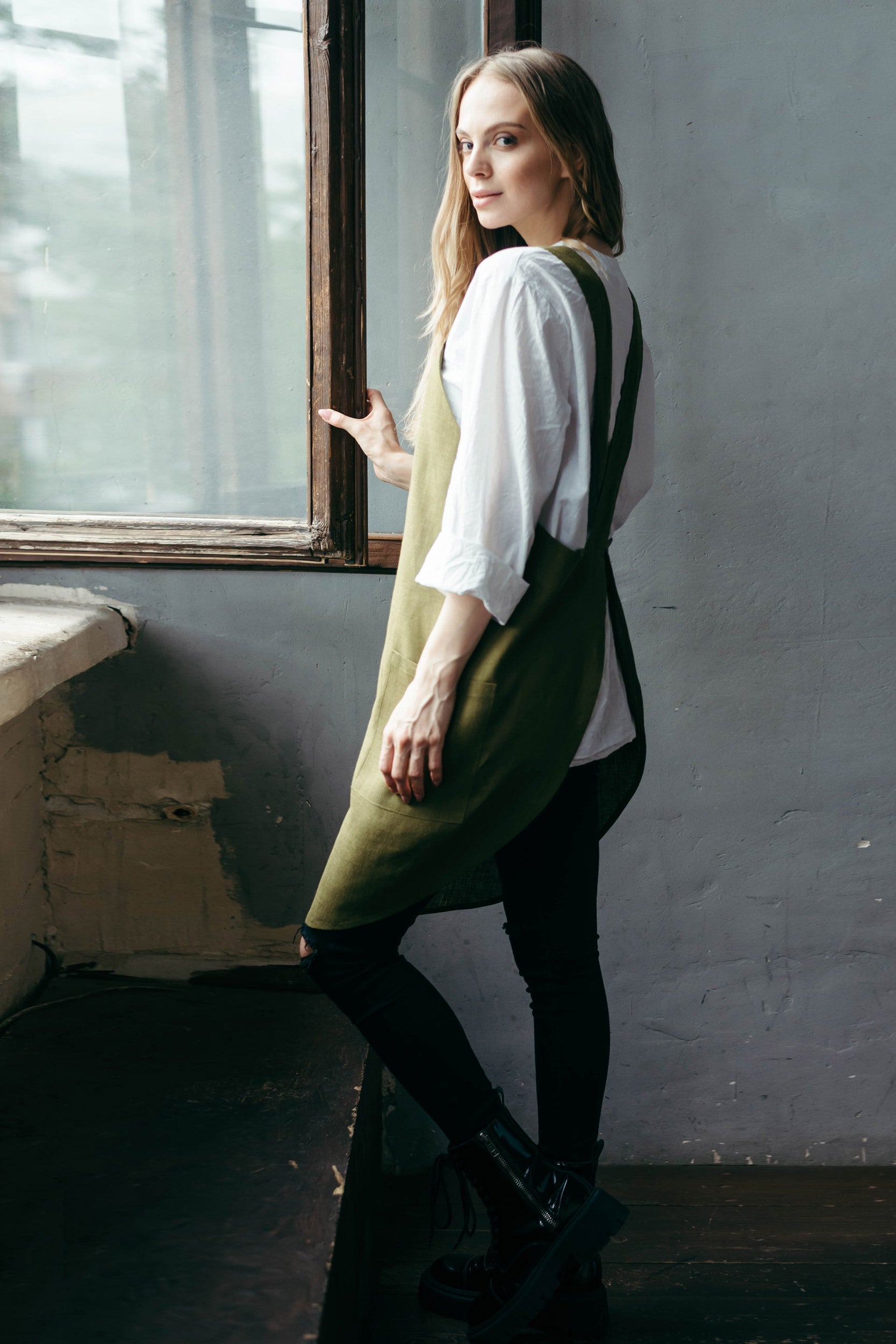 Woman wearing a green linen apron and white shirt against a gray wall.